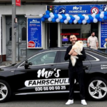 Mos Fahrschule Charlottenburg neue Filiale. Mo mit Hund am Arm und Auto im Vordergrund. Filiale im Hintergrund